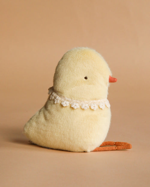 The Maileg Chicken Plush, a soft stuffed toy with a yellow body, orange beak and feet, and a white lace collar, sits on a beige surface against a plain beige background.
