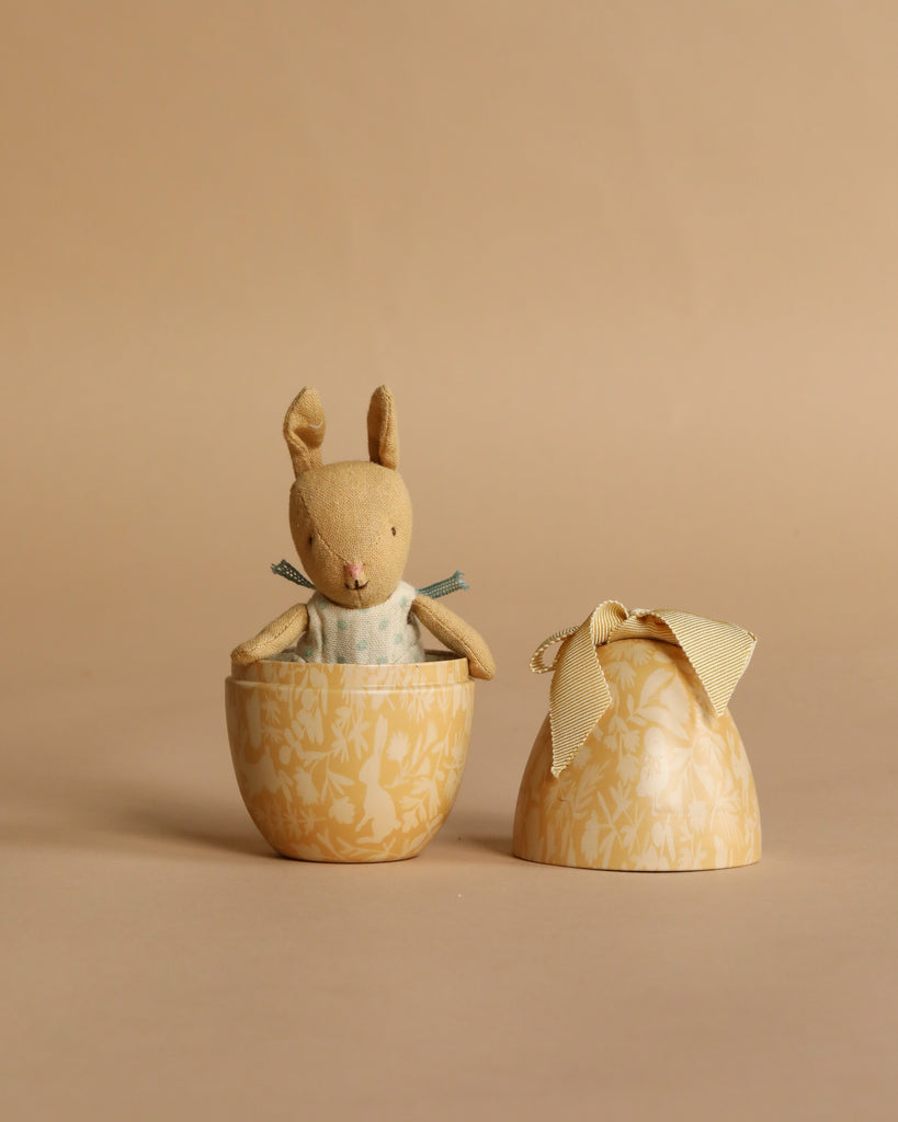The Maileg Easter Egg with Micro Rabbit, Small - Cream Yellow features a soft rabbit in a blue dress inside a yellow, floral-patterned egg-shaped container with a cream bow lid, set against a beige background.
