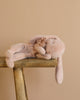 A soft Maileg Sleeping Bunny With Bed, crafted from recycled polyester, lies on its side on a rustic wooden shelf. The light pink plush toy features floppy ears and a decorative fabric flower near its head against a beige background.