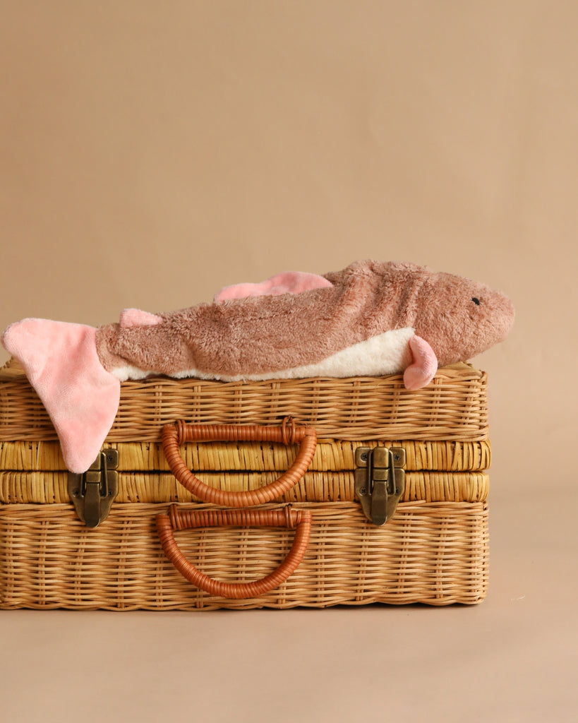A brown and pink Senger Naturwelt Cuddly Animal - Fish plush rests atop a closed wicker suitcase with leather handles and metal clasps, set against a plain beige background—a charming, sustainable soft toy for all ages.