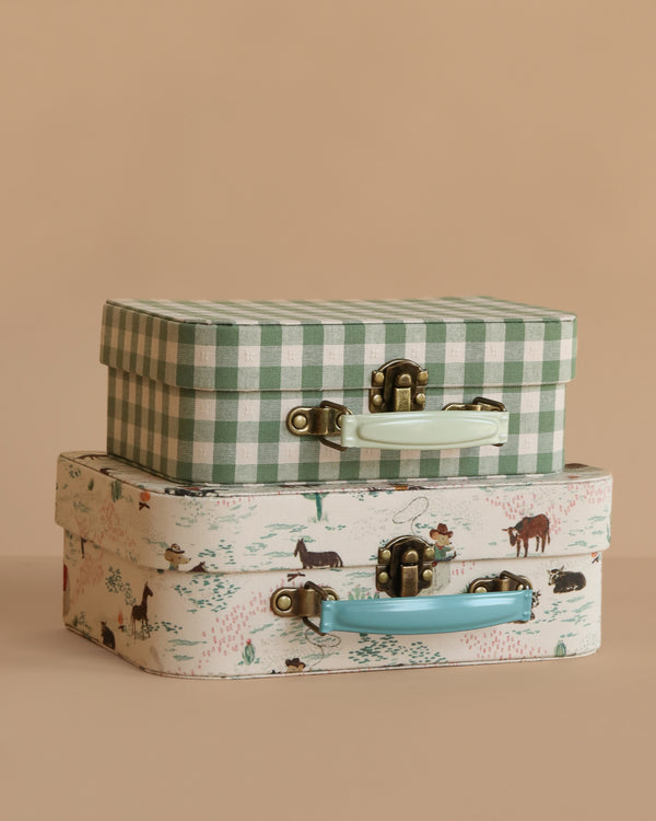 Two decorative suitcases are stacked—a green and white checkered toy suitcase with a white handle sits atop the Maileg Suitcase Set, 2-Pieces - Pony, featuring an animal and nature print with a blue handle, both against a beige background.
