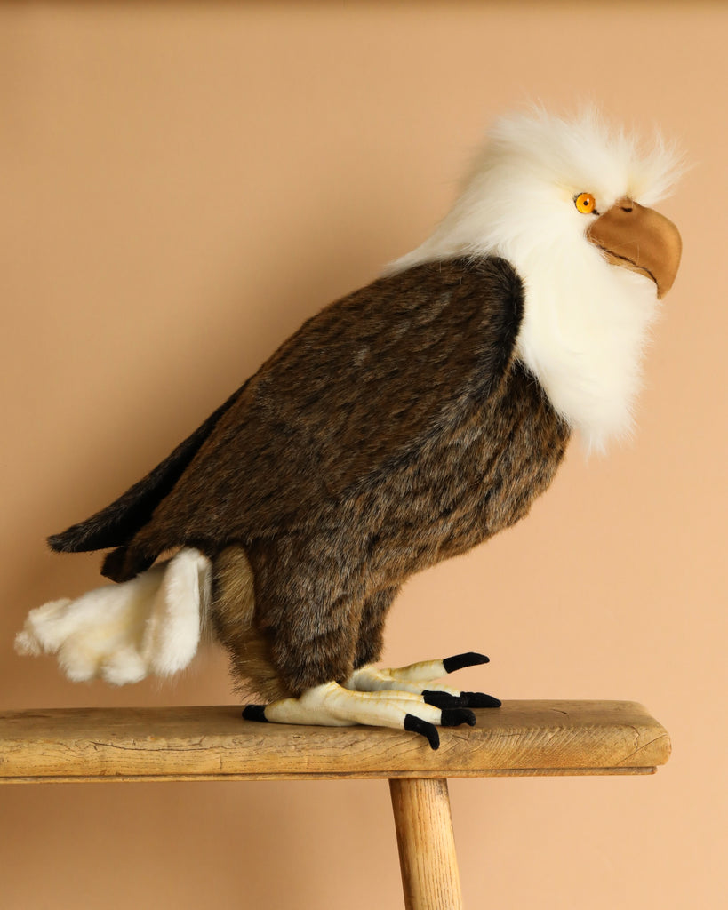 The American Eagle Stuffed Animal by HANSA Animals, with lifelike details like a white head, brown body, black talons, bright yellow eyes, and a prominent beak, is perched on a wooden stool against a beige background.