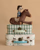 A Maileg Cowboy Set features a cowboy mouse on a plush rocking horse, displayed atop two Maileg suitcases—one green plaid, one with a pastoral print—arranged against a beige background.