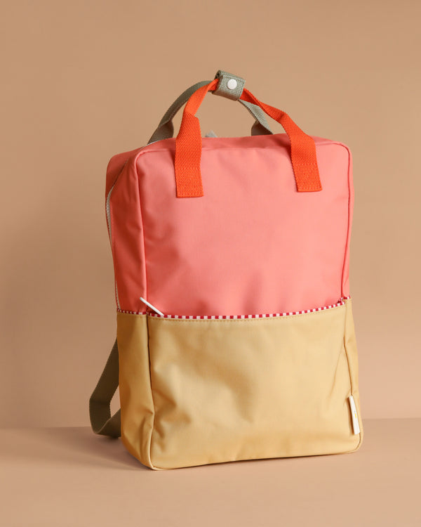 The Sticky Lemon Backpack Large | Color Blocking | Coral Pink + Reed Green features a two-tone design with coral pink and reed green, orange handles, beige straps, a white snap button on a gray loop, is made from recycled PET bottles, and has a waterproof lining.