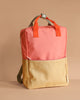 The Sticky Lemon Backpack Large | Color Blocking | Coral Pink + Reed Green features a two-tone design with coral pink and reed green, orange handles, beige straps, a white snap button on a gray loop, is made from recycled PET bottles, and has a waterproof lining.