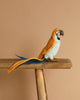 A Parrot Stuffed Animal, featuring artisanal craftsmanship and vibrant yellow, blue, and orange feathers, perches on a wooden beam against a beige background.