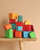 Grimm's Colored Waldorf Blocks, painted in vibrant orange, red, blue, green, and yellow hues, are stacked atop a wooden stool against a beige background. These non-toxic wooden blocks form a playful and balanced arrangement ideal for open-ended play.