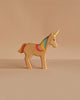 The Handmade Holzwald Unicorn Rainbow, a sustainable wooden toy with a yellow horn and rainbow mane, stands on a beige surface against a matching beige background.