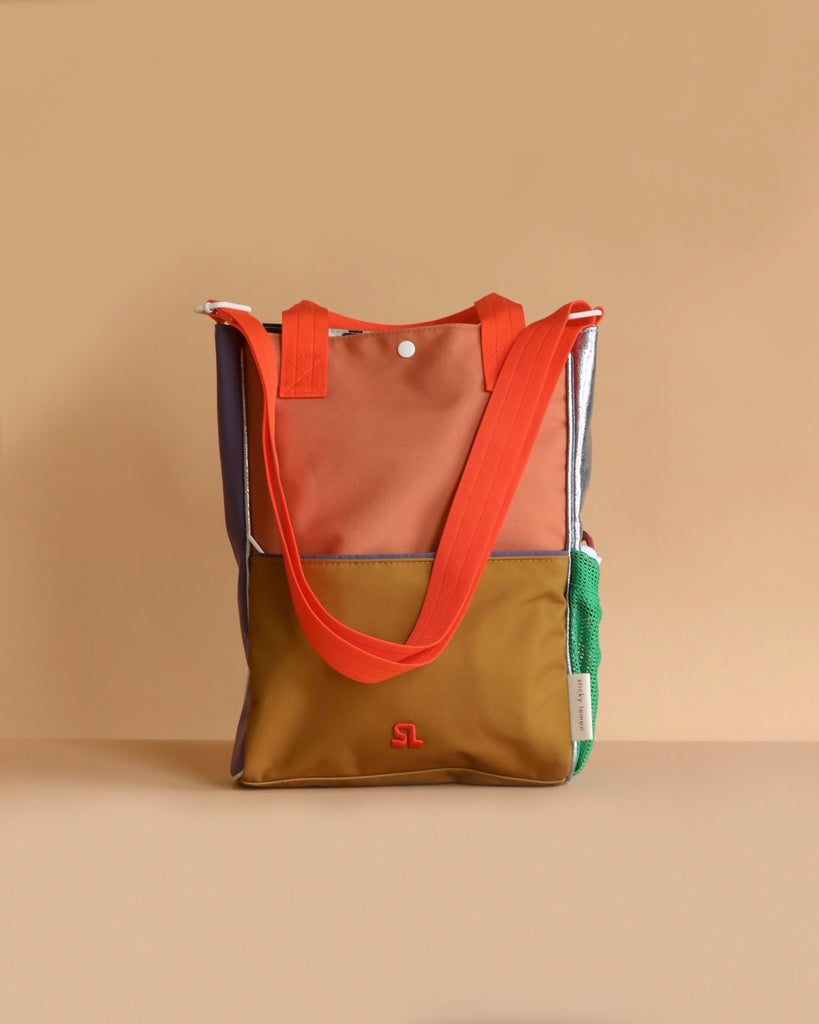 The Sticky Lemon Shoulder Bag | Leather Ball + Golden Cup features orange handles, a brown and tan front panel, green mesh side pocket, small white logo on the front—all crafted from recycled PET bottles—shown against a beige background.