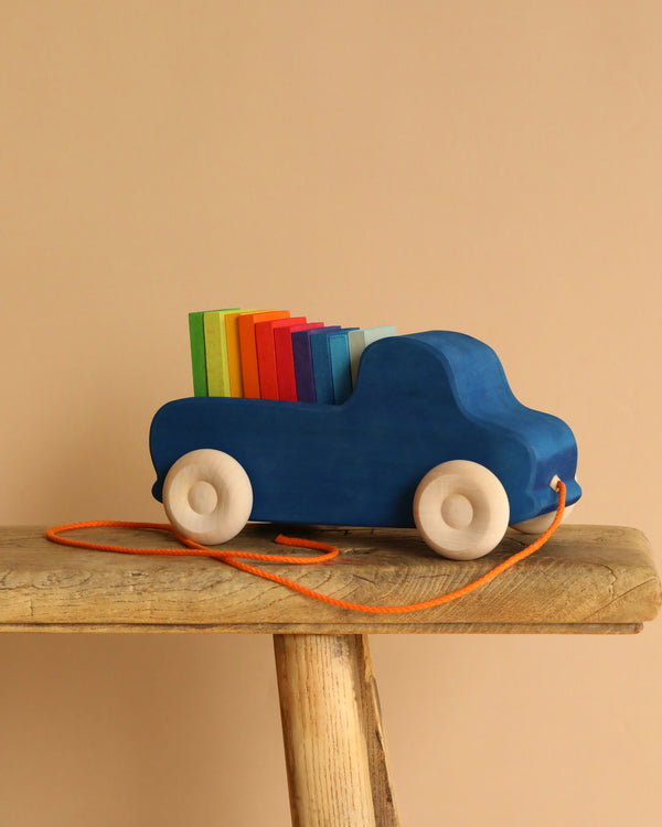 The Grimm's Pull Along Truck, a blue lime wood toy with white wheels and an orange string, rests on a wooden stool filled with colorful red, yellow, green, and blue blocks against a solid beige backdrop.