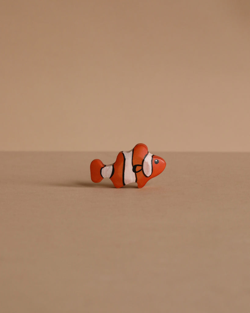 A small orange and white clownfish from the Handmade Holzwald Underwater World Set rests on a plain beige surface with a matching beige background.