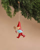 Hanging from a green Christmas tree branch against a beige background, the Maileg Metal Ornament, Pixy Spoon features a jolly elf adorned in red and blue, with a pointy hat and striped scarf. It showcases intricate handpainted details as it holds a wooden spoon.
