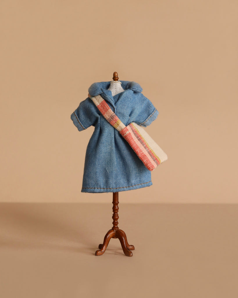The Maileg Mum Mouse Wardrobe Set features a miniature mannequin displaying a blue collared, short-sleeve coat and a cream shoulder bag with red and orange stripes, all set against a beige background.