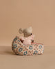 The Maileg Baby Mouse in Carry Cot - Rose features a small gray mouse in a pink sweater, resting inside a floral-patterned fabric carry cot, set against a beige background.