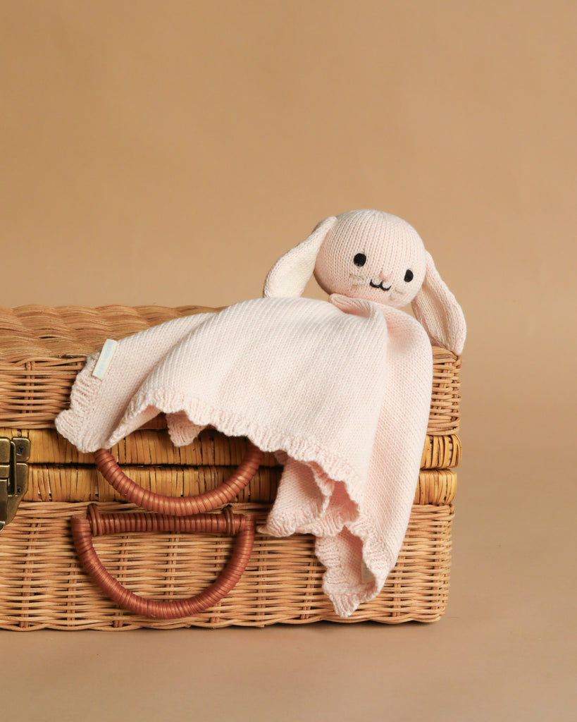 The Cuddle + Kind Organic Cotton Baby Bunny Lovey (Blush), a soft hand-knit pink bunny with floppy ears, rests on a closed wicker basket with two handles against a plain beige background.