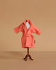 A small mannequin displays the Maileg Extra Clothing: Bathrobe - Coral, a textured pink cotton robe with a belt, set against a plain beige background.