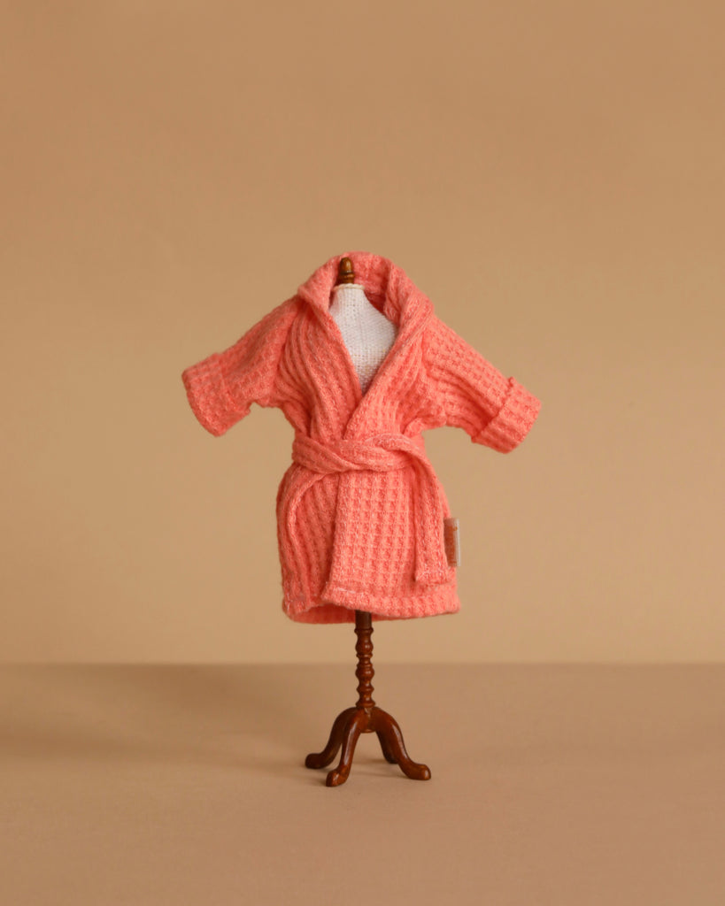 A small mannequin displays the Maileg Extra Clothing: Bathrobe - Coral, a textured pink cotton robe with a belt, set against a plain beige background.