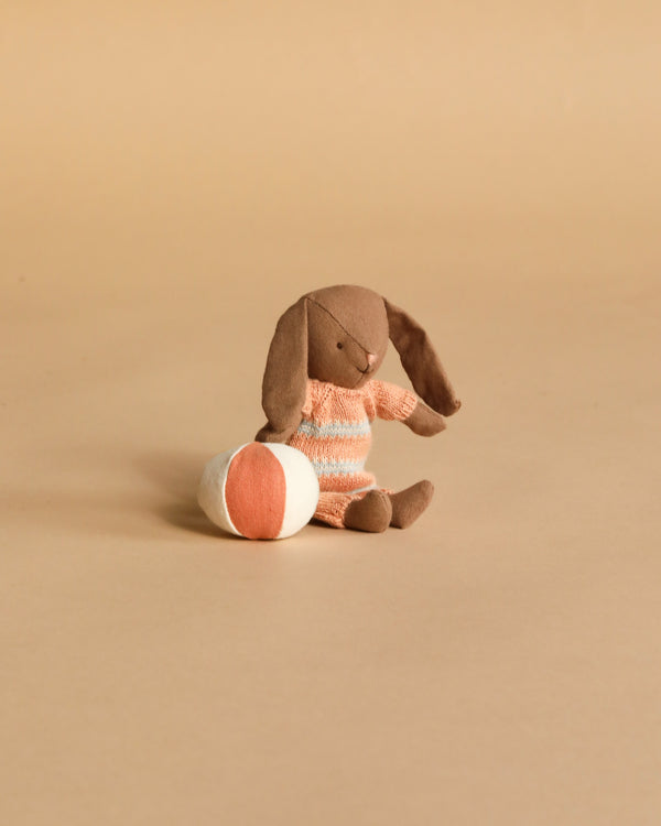 The Maileg Micro Bunny With Ball - Brown - Dusty Rose (Magnetic Hands) sits on a beige surface beside a round orange and white ball, making it a charming storytelling toy.