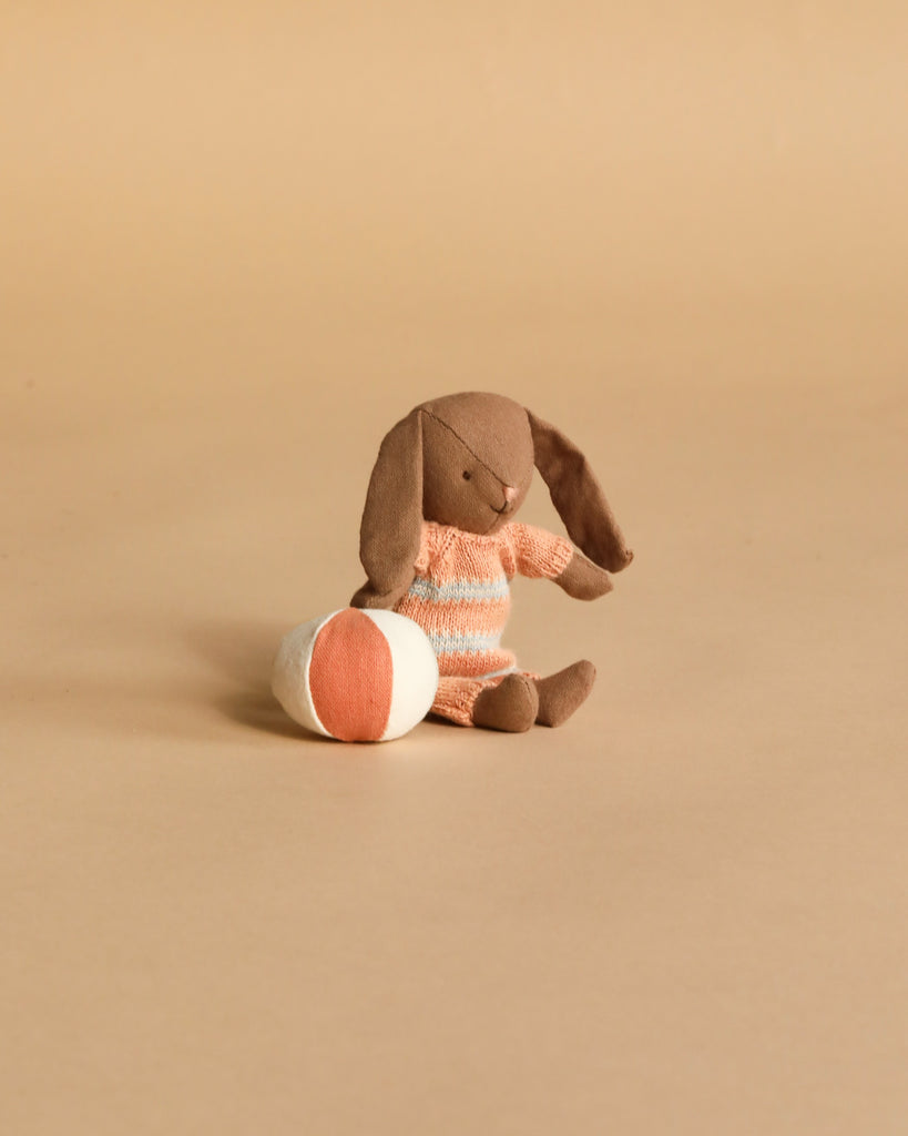 The Maileg Micro Bunny With Ball - Brown - Dusty Rose (Magnetic Hands) sits on a beige surface beside a round orange and white ball, making it a charming storytelling toy.