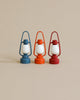 Three Maileg Lanterns are placed side by side against a neutral background. Each non-rechargeable lantern features a white globe and comes in a different color: blue, orange, and red. The lanterns are perfectly aligned, creating a visually appealing arrangement.