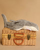 The Senger Naturwelt Cuddly Animal - Shark, a small gray plush with a white underside and soft teeth, rests atop a closed wicker suitcase with leather handles and brass clasps against a beige background.