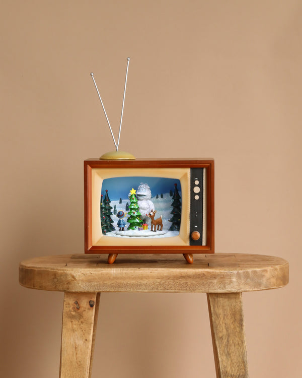 The Christmas Music TV - Yeti and Rudolf features a vintage light-up TV with antennas on a wooden table, showcasing snowy trees, a snowman, and Rudolf on the screen—an ideal holiday decoration against a plain beige background.