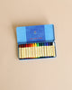 A tin case of sixteen colourful, non-toxic Stockmar wax stick crayons neatly arranged by color spectrum in a blue cardboard box with a decorative lid, placed on a light brown surface.