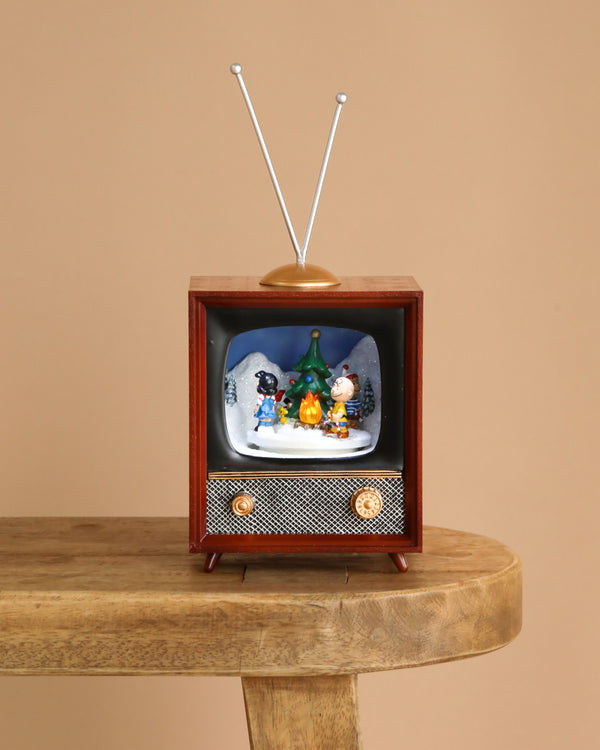 The Christmas Music TV - Charlie Brown sits on a wooden table, featuring a vintage design with metal antennae and golden knobs. Inside the screen, cartoon figures gather around a tree and campfire in a festive winter scene.