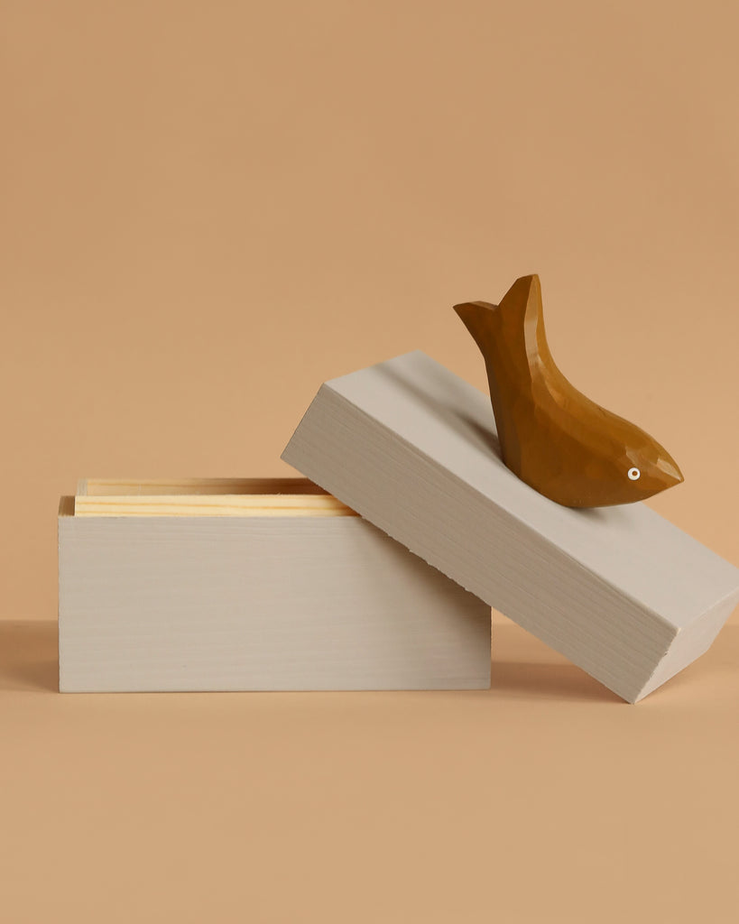 The Ferm Living Hand Carved Wooden Harbour Box - Fish, a beige rectangular box with a partially open lid, sits on a tan surface. A bird figurine made from FSC certified aspen wood rests atop the lid, ideal for kids' room decor against a light brown background.