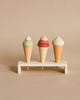 Three handmade ice cream cone toys with green, pink, and white swirls on top, displayed on a tiny wooden shelf against a neutral background.