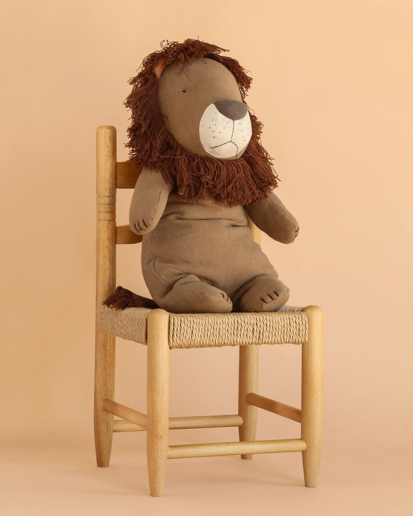 The Maileg Safari Friends - Big Lion, a soft handmade brown lion plush with a shaggy mane, sits upright on a woven wooden chair against a beige background.