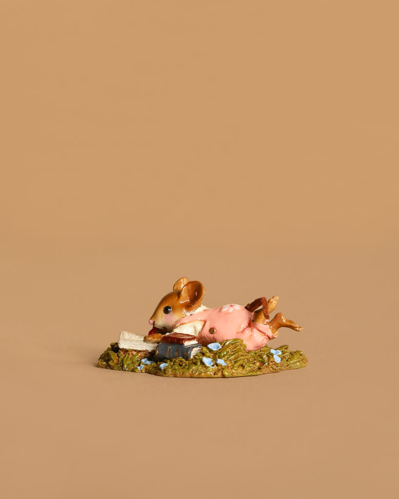 The Wee Forest Folk Tiny Collectible - Meadow Musing features a mouse in a pink dress lying on green grass among blue flowers, reading a book. This charming handmade figurine is beautifully displayed against a tan background. Not a toy.