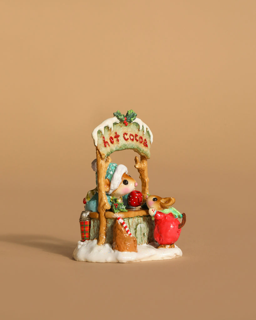 The Wee Forest Folk Tiny Collectible - Hot Cocoa Comin' Up! (Not a Toy) features two hand-sculpted mice at a snowy cocoa stand, surrounded by candy canes, gifts, and holiday decor—one serving cocoa and one in a red coat sipping.