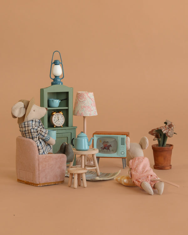 Two toy mice relax in a cozy Maileg scene featuring the Maileg Cozy Evening with Granddad Set, complete with a lamp, TV, potted plant, clock, and stylish furniture—all displayed on a beige background for charming miniature decor.