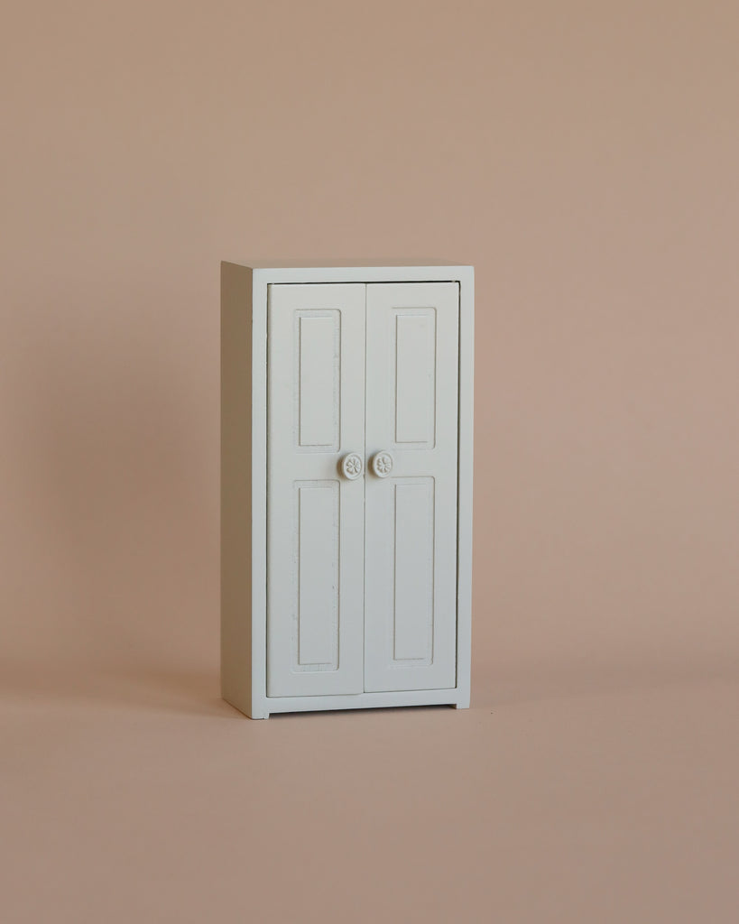 The Maileg Mum Mouse Wardrobe Set, a compact white closet with double doors and round knobs for storing mice clothes, stands on a plain beige surface with a beige background.