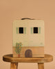 The Maileg Suitcasa Furnished Set—a suitcase designed to resemble a charming house, featuring two windows with green shutters, a door, a potted plant, and "Suit Casa" written on the front in whimsical Maileg style—is displayed on a wooden stool against a beige background.