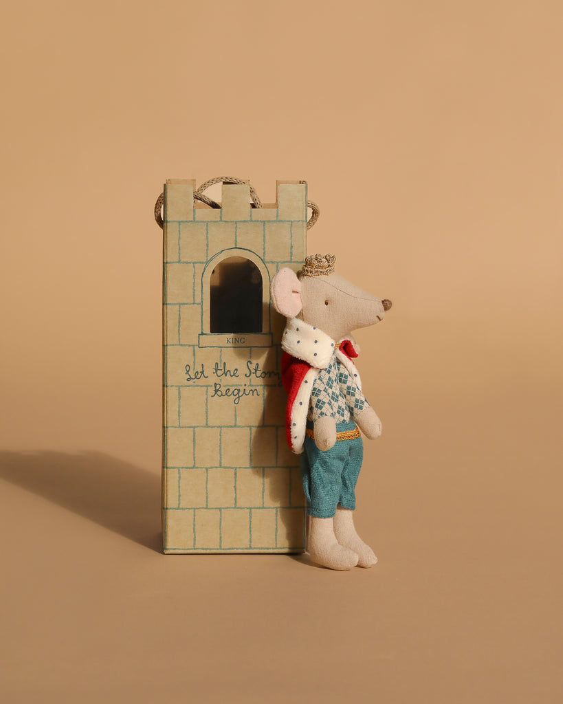 A Maileg Fairytale Set is displayed with a mouse dressed as a king leaning against a charming castle tower, which features a window and the phrase “Let the Story begin.” Beige background.