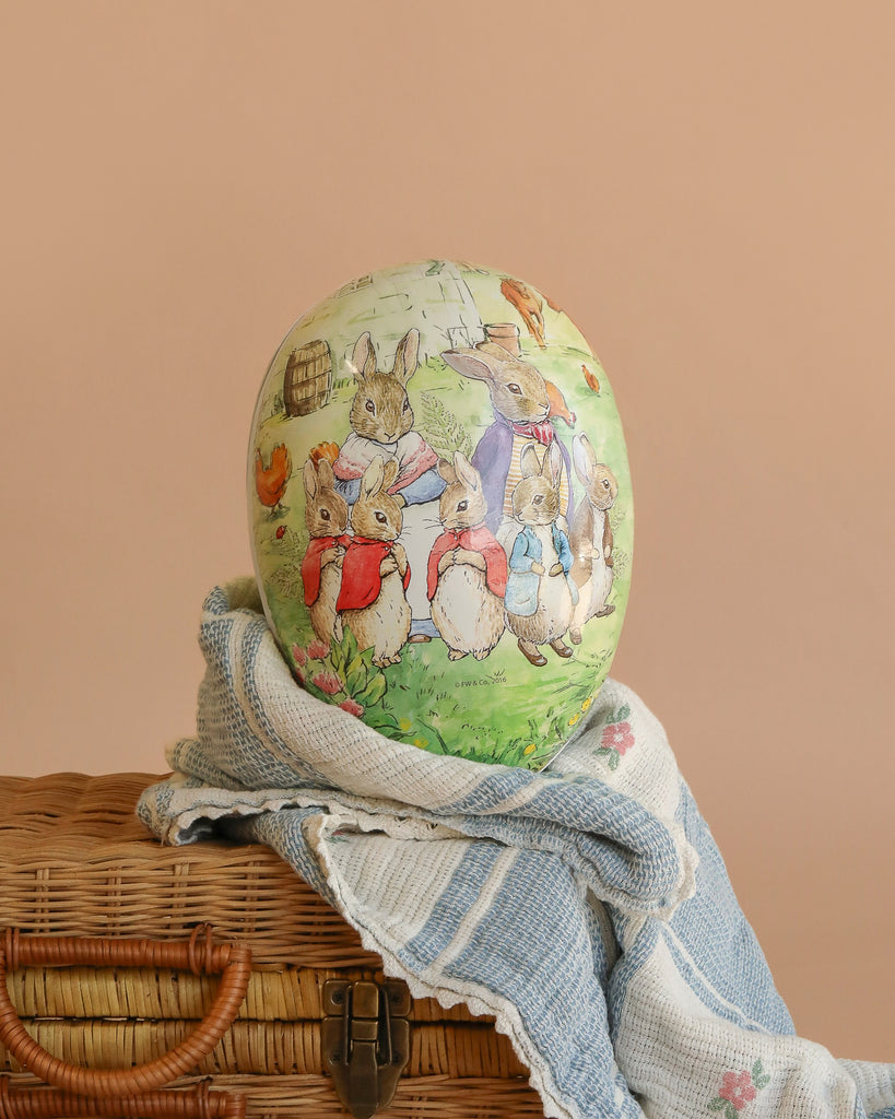 The Cardboard Beatrix Potter Egg, Extra Large (Rabbit Family In The Garden) features clothed rabbits by a stone wall in a garden, resting on a woven basket with a blue and white blanket, reminiscent of handcrafted Nestler GmbH eggs.