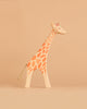 A handcrafted wooden giraffe figurine from the Ostheimer Handmade Wooden Safari Animals Set, featuring orange spots, stands on a plain beige surface against a matching beige background, reflecting the timeless quality and sustainable materials characteristic of Ostheimer toys.
