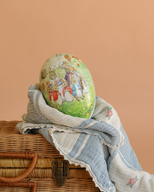 A large Cardboard Beatrix Potter Egg featuring the Rabbit Family in the Garden rests on a blue and white textured cloth atop a woven wicker basket, set against a plain peach background.