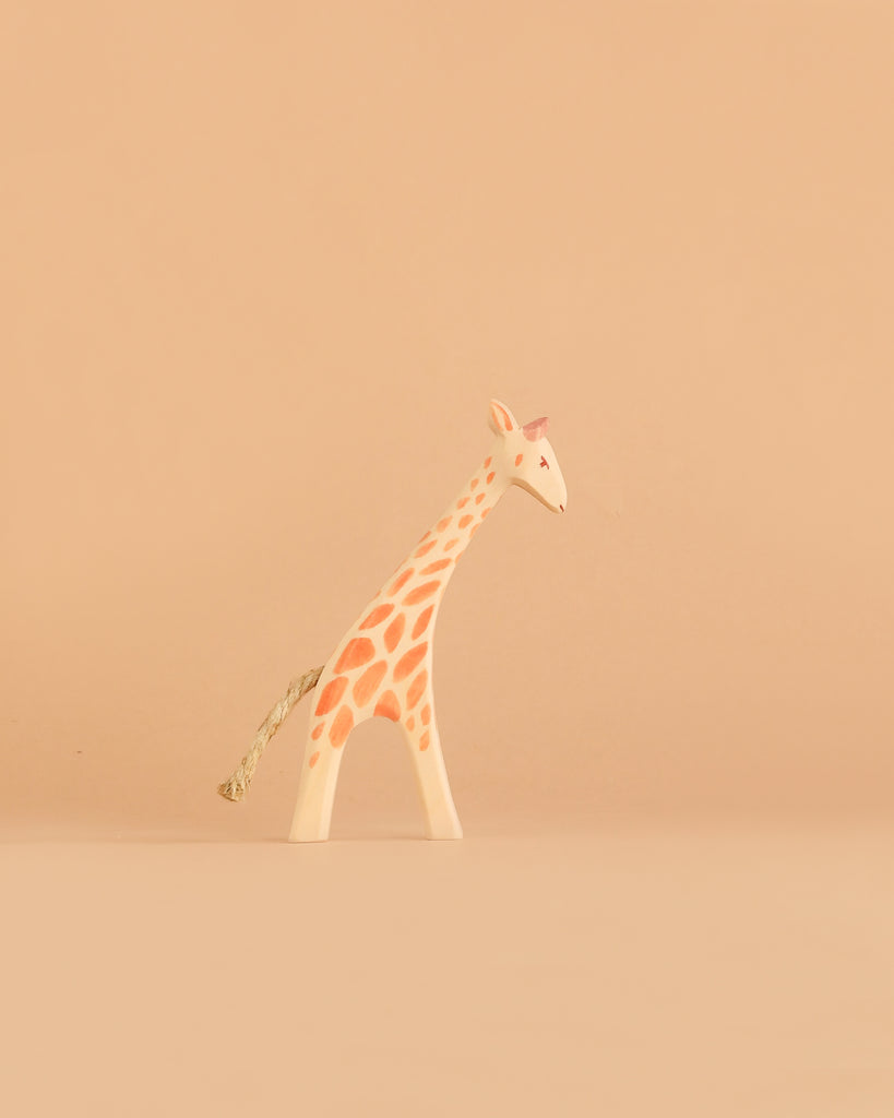 A small, minimalist giraffe figurine with orange spots and a rope tail stands on a plain light brown background, inspired by the timeless charm of the Ostheimer Handmade Wooden Safari Animals Set and crafted from sustainably sourced wood.