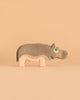 One piece from the Ostheimer Handmade Wooden Safari Animals Set—a handcrafted wooden hippopotamus inspired by classic Ostheimer toys and painted in soft gray and pink—stands on a beige surface with a matching background. Made from sustainably sourced wood, this delightful figure blends durability with timeless design.