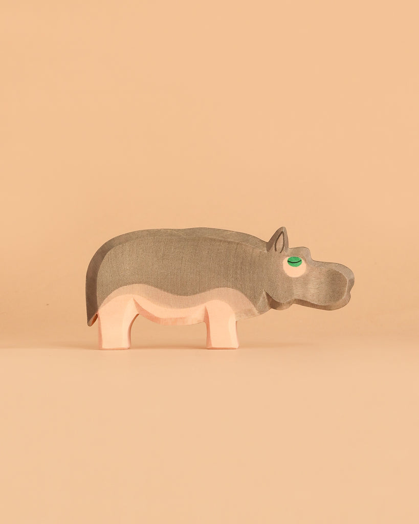 One piece from the Ostheimer Handmade Wooden Safari Animals Set—a handcrafted wooden hippopotamus inspired by classic Ostheimer toys and painted in soft gray and pink—stands on a beige surface with a matching background. Made from sustainably sourced wood, this delightful figure blends durability with timeless design.