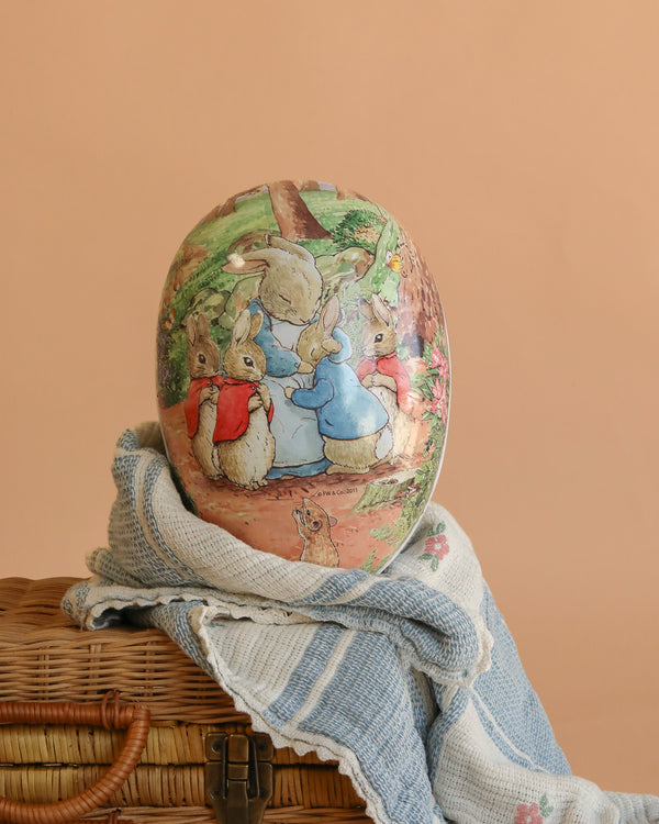 The Cardboard Beatrix Potter Egg, Extra Large (Mother Rabbit with Three Baby Rabbits), handcrafted in Germany, features a mother rabbit and her three babies and is displayed on a blue-and-white cloth over a wicker basket.