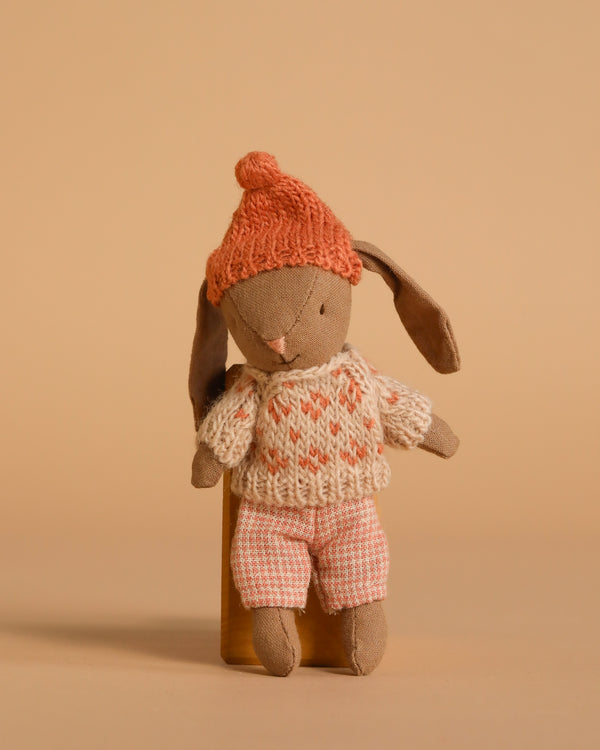 The Maileg Winter Bunny, Micro - Brown - Rose wears a knitted orange hat, cream and orange sweater, and pink checkered shorts—ideal for miniature dollhouse play or pairing with Maileg accessories against a beige background.