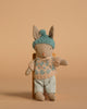 The Maileg Winter Rabbit, Micro - Sand - Mint wears a blue knit hat, beige and blue sweater, and light blue pants, sitting on a wooden chair against a beige background—ideal for dollhouse displays or Maileg accessories.