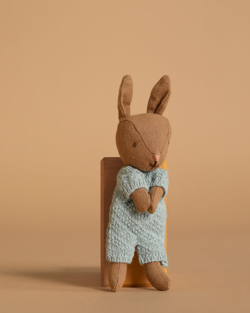 The Maileg Rabbit Micro - Brown wears a blue knitted suit and sits upright against a wooden block, ideal for miniature dollhouses or pairing with other Maileg accessories, displayed on a plain beige background.