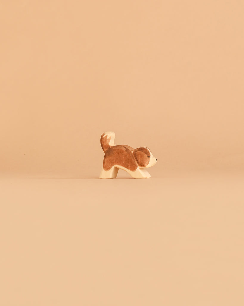 A small, brown and white dog figurine from the Ostheimer Handmade Wooden Dog Family Set, crafted from sustainably sourced wood in the signature Ostheimer style, stands on a plain beige background.