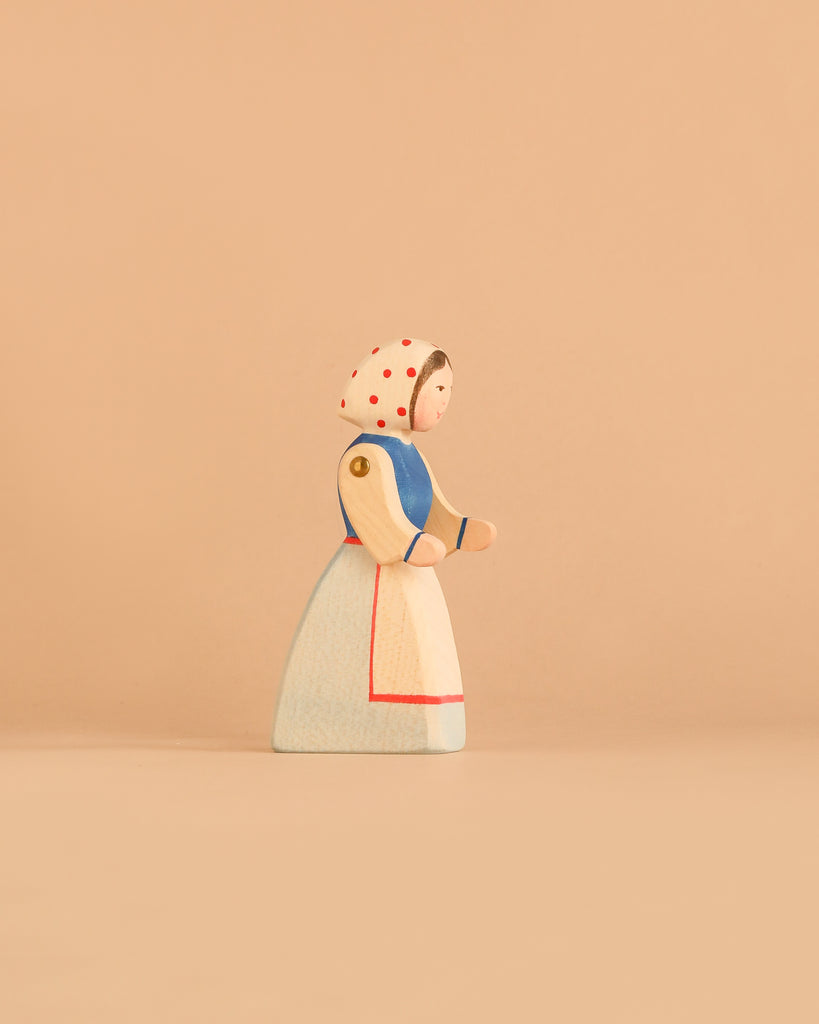 Introducing the Ostheimer Handmade Wooden Swan Girl Set: a beautifully handcrafted wooden figurine of a woman in a bonnet with red dots, a blue shirt, and a long white skirt, standing on a beige background. Inspired by Ostheimer toys and carefully made from sustainably sourced wood, this set captures the timeless charm of traditional craftsmanship.
