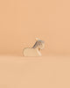 A small, minimalist goat figurine from the Ostheimer Handmade Wooden Farm Animals Set, handcrafted from sustainably sourced wood with grey and white accents, sits on a light peach background.